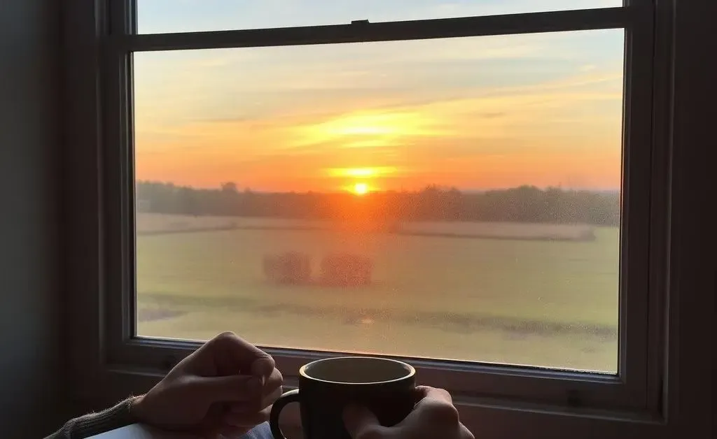 A person enjoying morning coffee while journaling, with a peaceful sunrise visible outside the window.